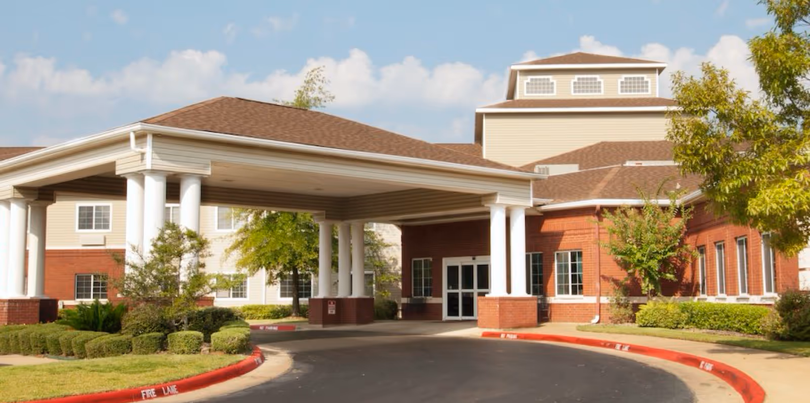 Exterior view of Garden Estates of Tyler senior living facility showing the entrance with a covered driveway supported by white columns, surrounded by landscaped bushes and trees under a partly cloudy sky.