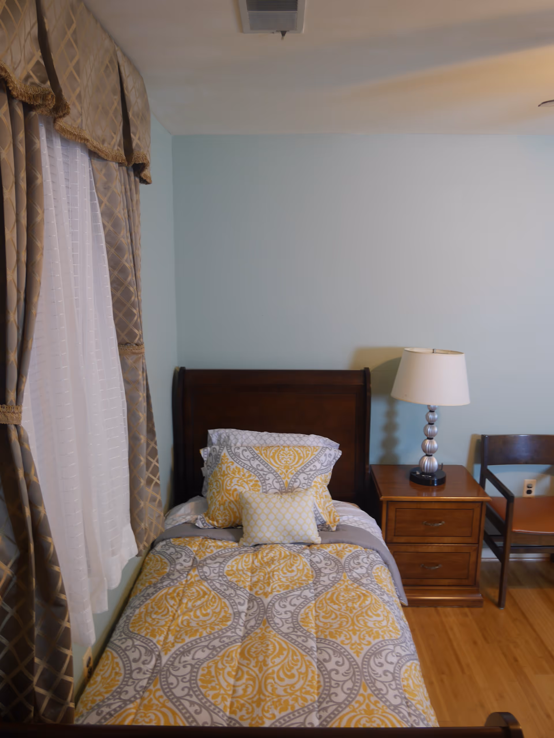 Single bed with yellow and gray patterned bedding next to a wooden nightstand, lamp, and curtained window in a tidy bedroom.