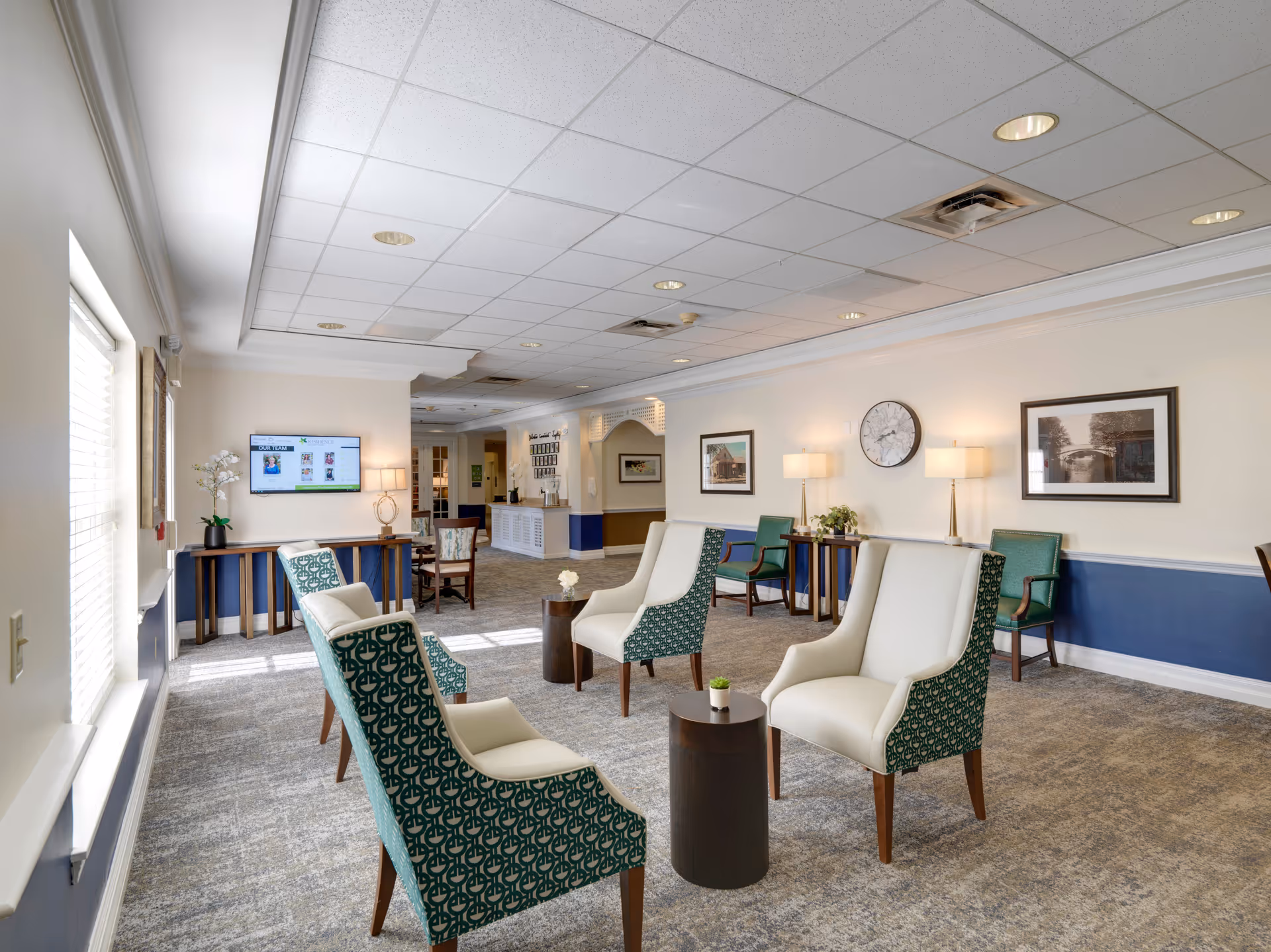 A bright and spacious senior living facility common area with patterned armchairs and white cushioned chairs arranged around small round tables. The room has a carpeted floor, white walls with blue wainscoting, framed artwork, a wall clock, and several lamps providing warm lighting. A flat-screen TV is mounted on the wall near a window with blinds, and there is a reception desk visible in the background.