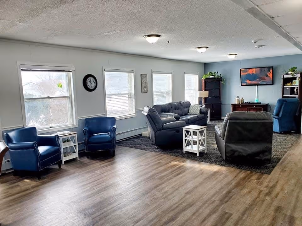 Spacious communal living room with sofas, recliners, blue armchairs, side tables, and a wall-mounted TV near several windows.