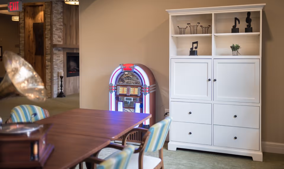 Interior view of a room with a wooden table surrounded by striped cushioned chairs. A vintage jukebox with colorful lights is placed against a beige wall next to a white cabinet with shelves displaying decorative musical note sculptures and small plants. In the background, there is a hallway with a visible exit sign and a fireplace.