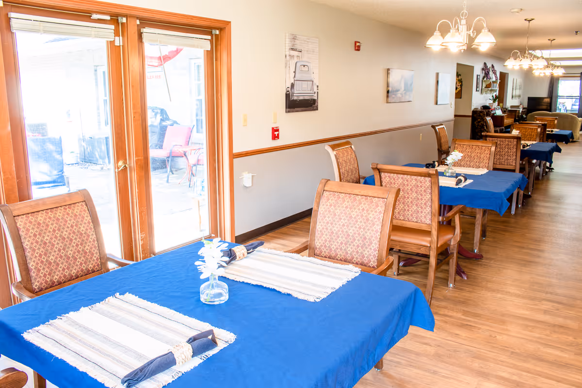 A bright dining area with blue-covered tables and wooden chairs lined along a long room with glass doors on the left.