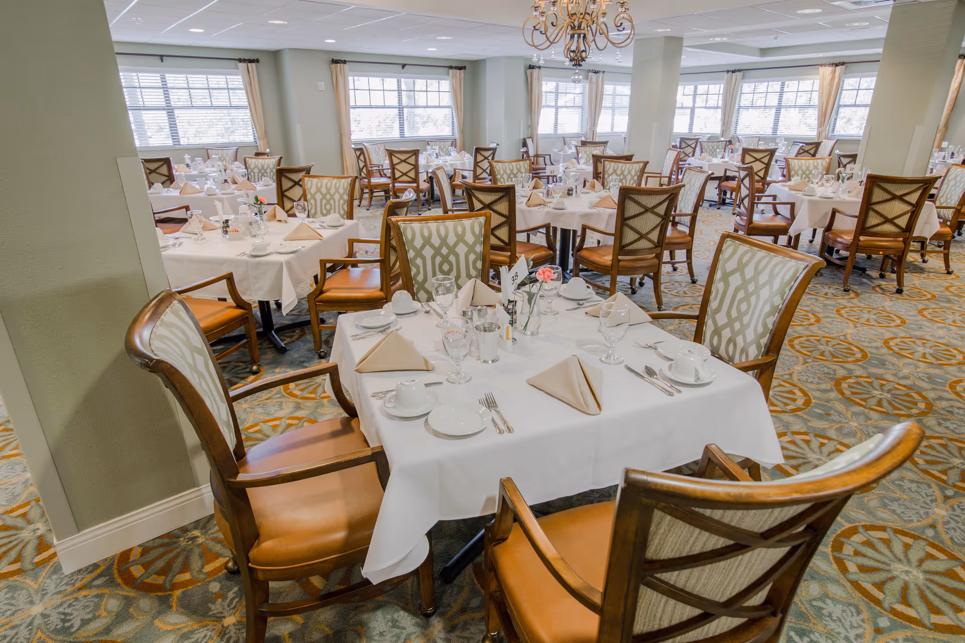A spacious dining room with multiple tables covered in white tablecloths, each set with beige napkins, glassware, cups, and silverware. The room features patterned carpet, large windows with beige curtains, and wooden chairs with cushioned seats and backs. A chandelier hangs from the ceiling.