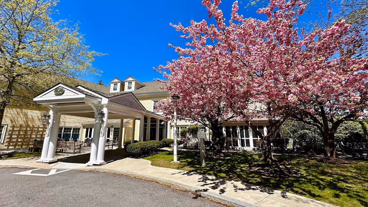 Exterior view of Orchard Hill at Sudbury facility with a covered entrance, yellow siding, and blooming pink cherry blossom trees under a clear blue sky.