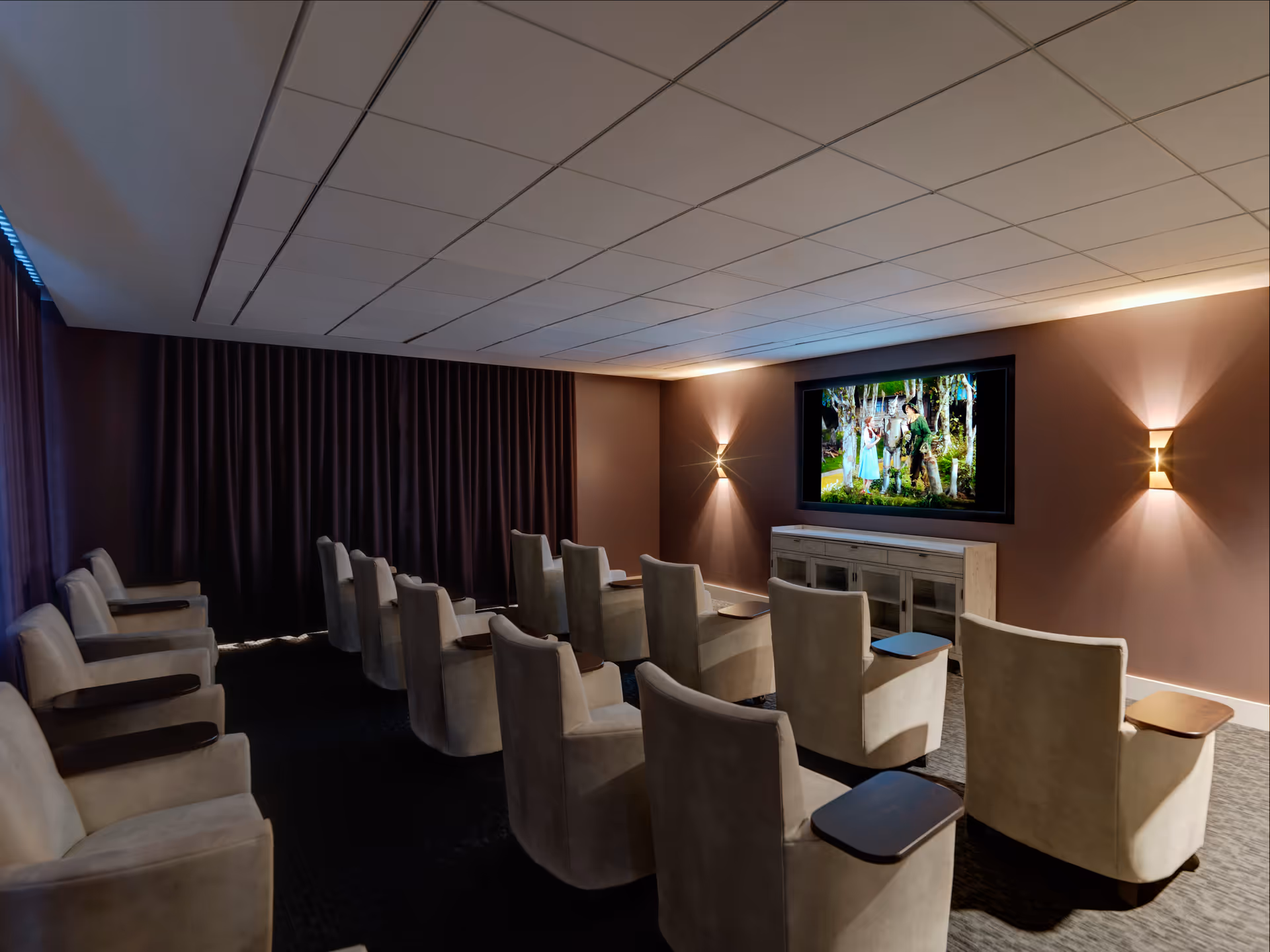 A small theater room with three rows of beige armchairs, each equipped with a wooden armrest tray, facing a large screen showing a scene from a movie. The room has dark curtains on one side and soft wall lighting on the other.