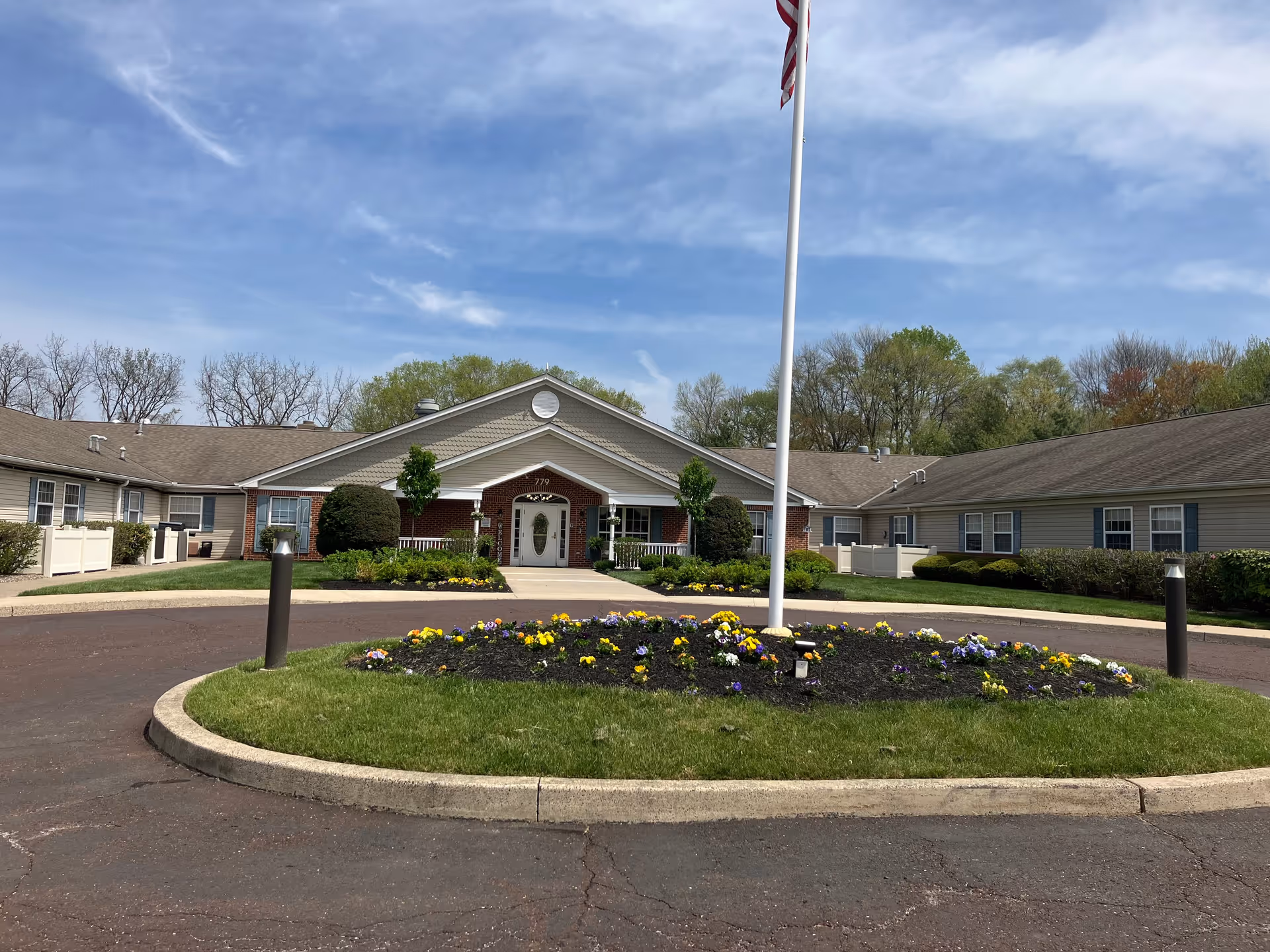 Front exterior view of Arden Courts - ProMedica Memory Care Community in Warminster, showing a single-story building with a peaked roof, a circular driveway with a flower bed in the center, and an American flag on a flagpole.