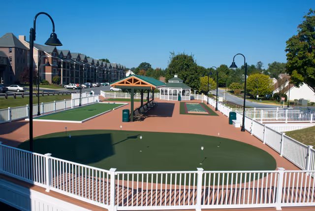 Outdoor recreational area at Phoebe Berks featuring a putting green, shuffleboard court, benches under a shaded pavilion, white fencing, and a gazebo in the background, surrounded by trees and residential buildings.