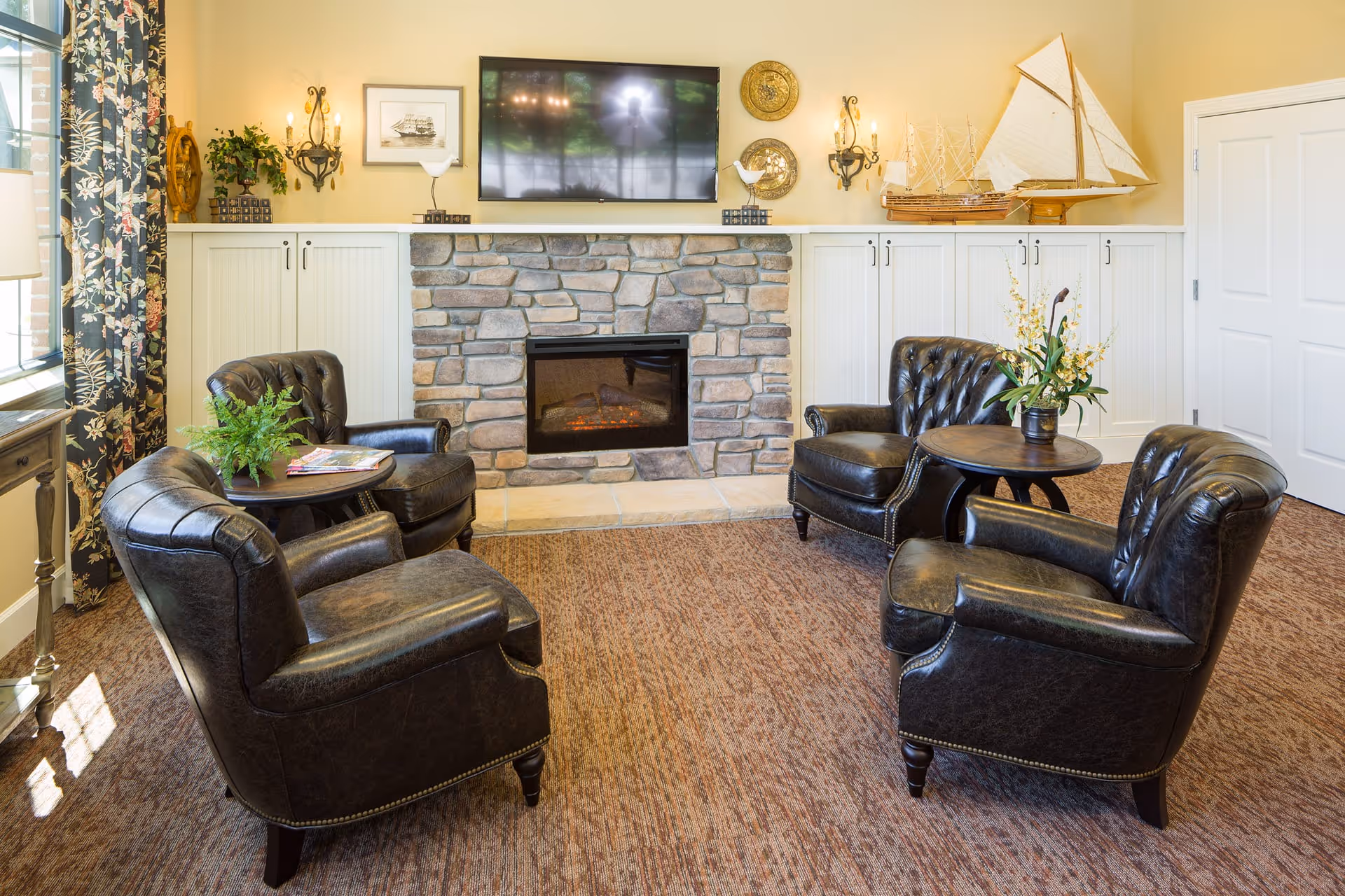Cozy sitting area with four leather armchairs around small tables facing a stone fireplace topped by a wall-mounted TV and decorative shelving.
