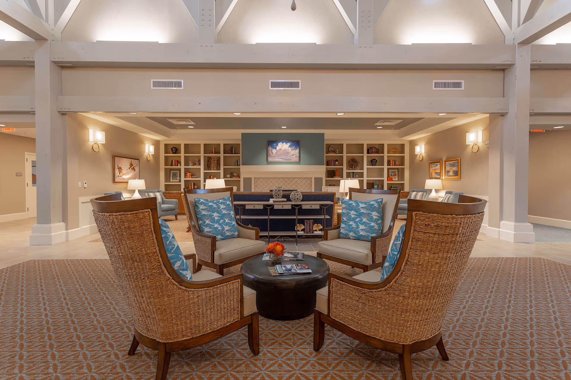 Comfortable lounge area with wicker armchairs and blue patterned pillows arranged around a round coffee table in a well-lit common room with built-in shelves and a fireplace.