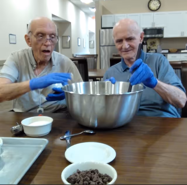 Two elderly men wearing blue gloves stirring ingredients in a large metal bowl at a communal dining table with a kitchen area in the background.