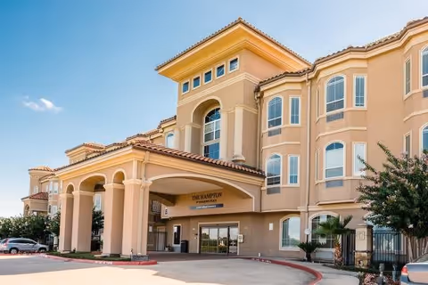 Exterior view of The Hampton at Meadows Place senior living facility showing a multi-story building with beige walls, large windows, and a covered entrance with columns. There are some trees and parked cars visible near the entrance under a clear blue sky.
