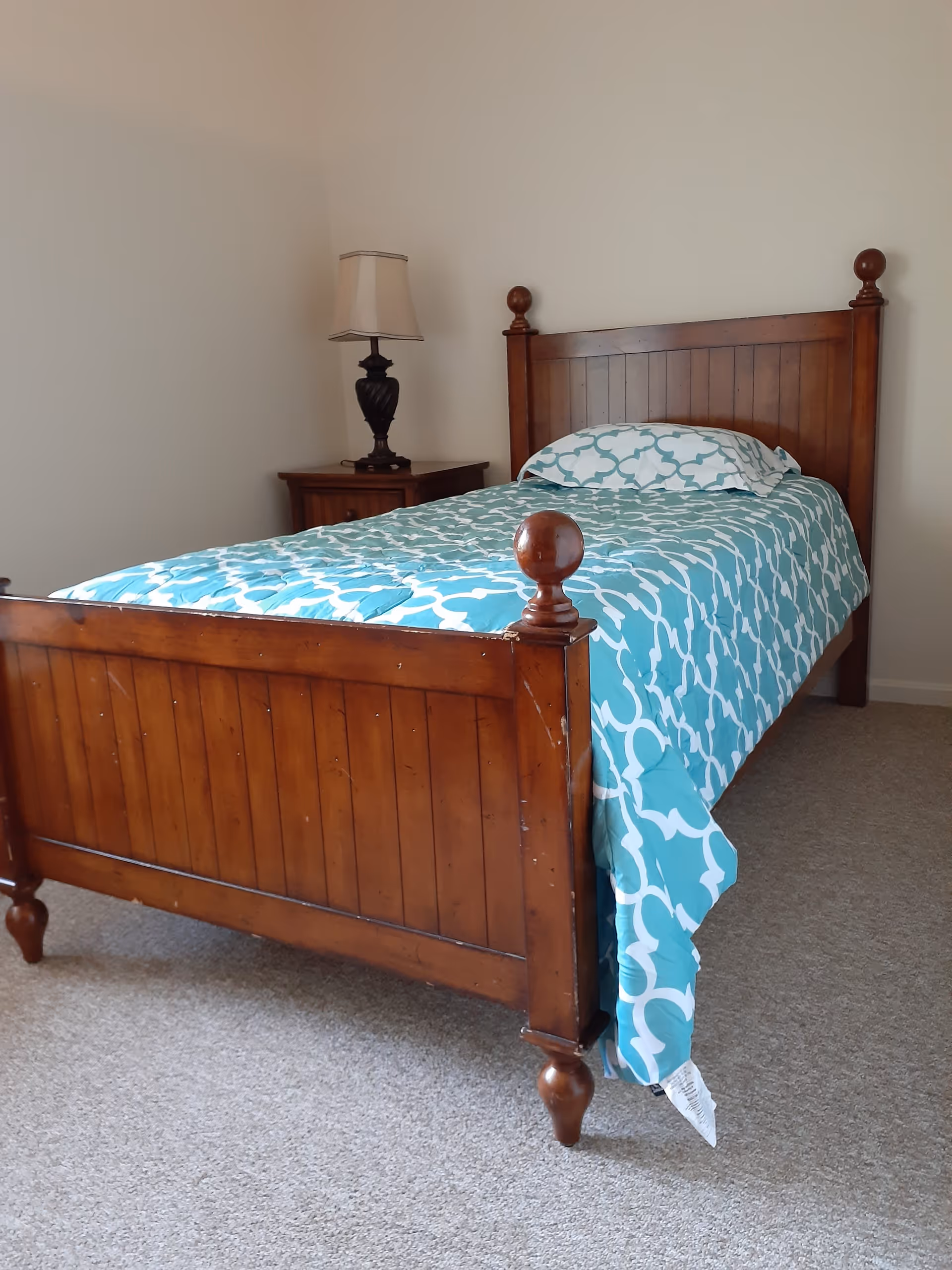 A simple bedroom with a wooden single bed featuring a blue and white patterned bedspread and matching pillow. Next to the bed is a wooden nightstand with a dark-colored lamp on it. The room has beige carpet and plain white walls.