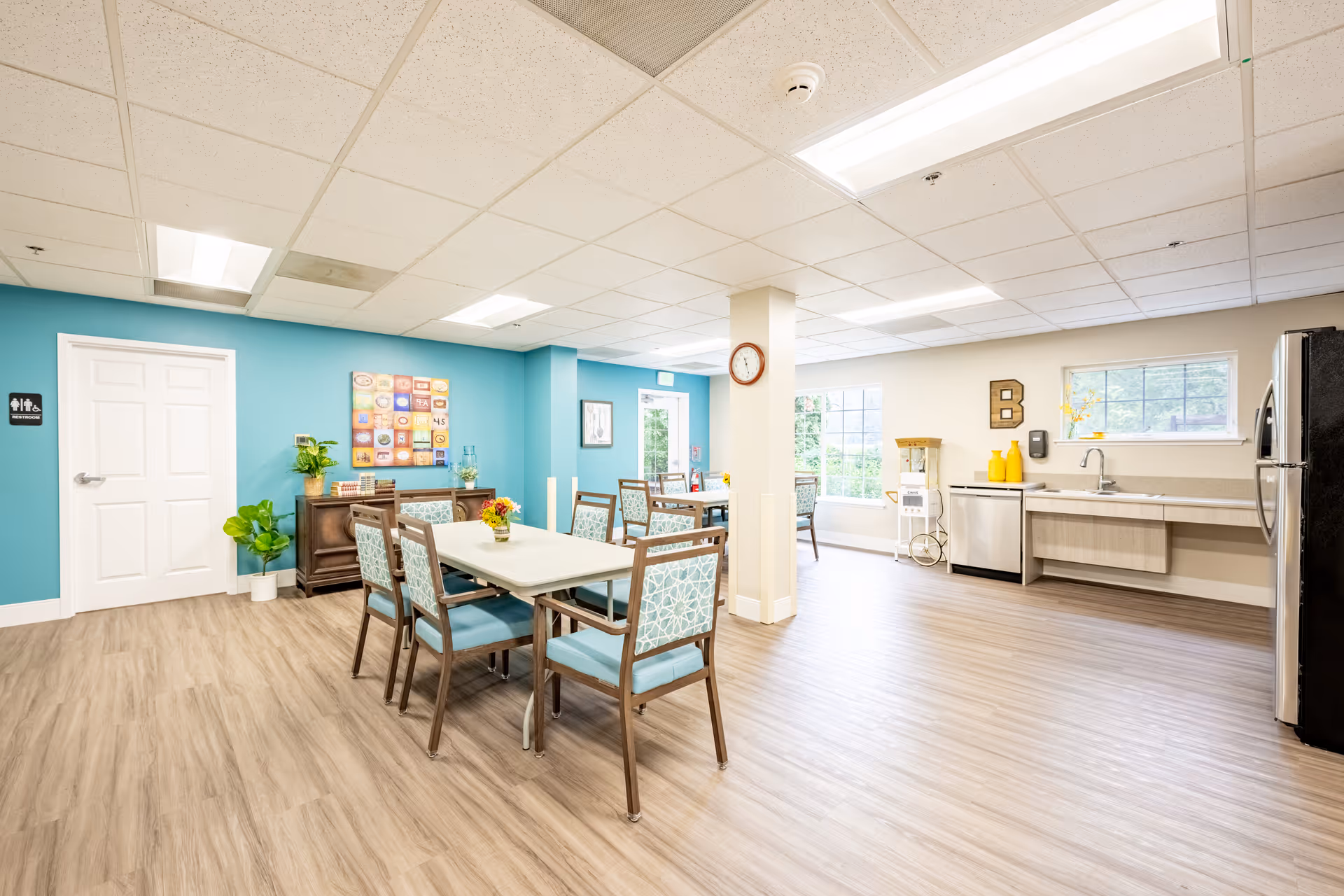 A bright and spacious dining area in a senior living facility with light wood flooring, teal accent walls, and multiple tables with chairs. There is a kitchenette area with a sink, dishwasher, and refrigerator. Large windows allow natural light to fill the room. A clock is mounted on a central pillar, and decorative plants and artwork adorn the space.