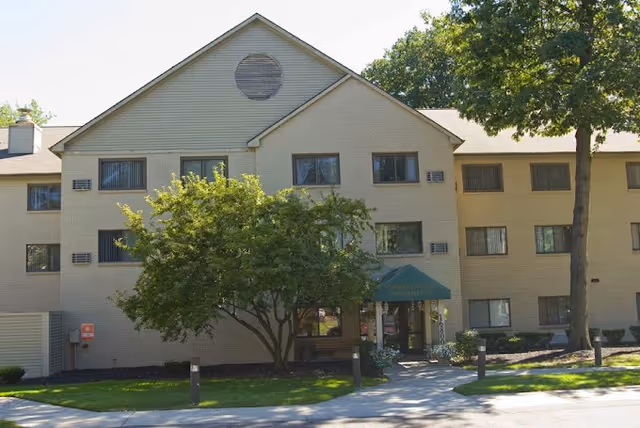 Exterior view of a three-story beige brick apartment building with multiple windows, a green awning over the entrance, and trees and landscaping in front.