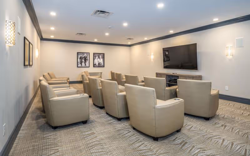 A small theater-style room with two rows of beige recliner chairs facing a large flat-screen TV mounted on the wall. The room has neutral-colored walls with modern wall sconces and framed black-and-white photos on the far wall. The carpet has a geometric pattern, and the ceiling has recessed lighting.