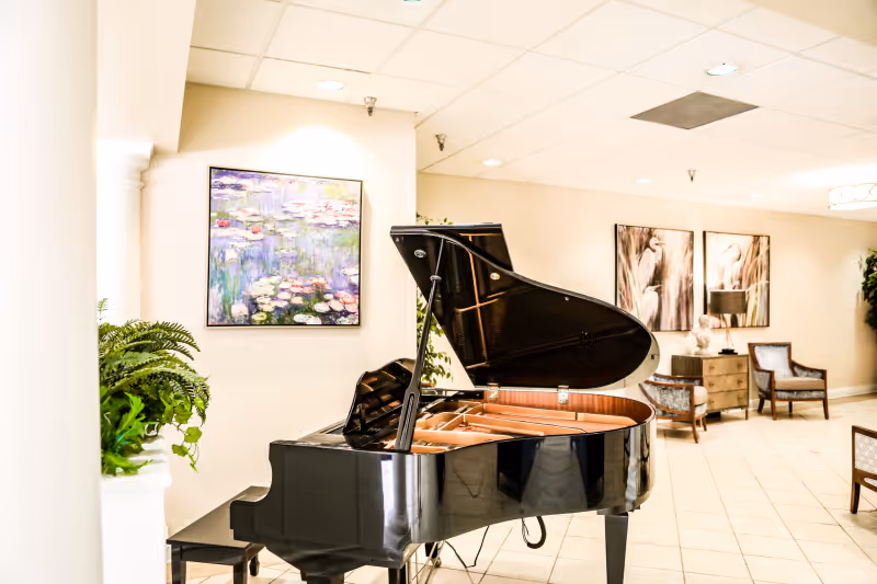 Interior view of a common area in a nursing home featuring a black grand piano with its lid open, a bench, framed artwork on the walls, two armchairs, a small chest of drawers with a lamp and decorative bust, and some green plants.
