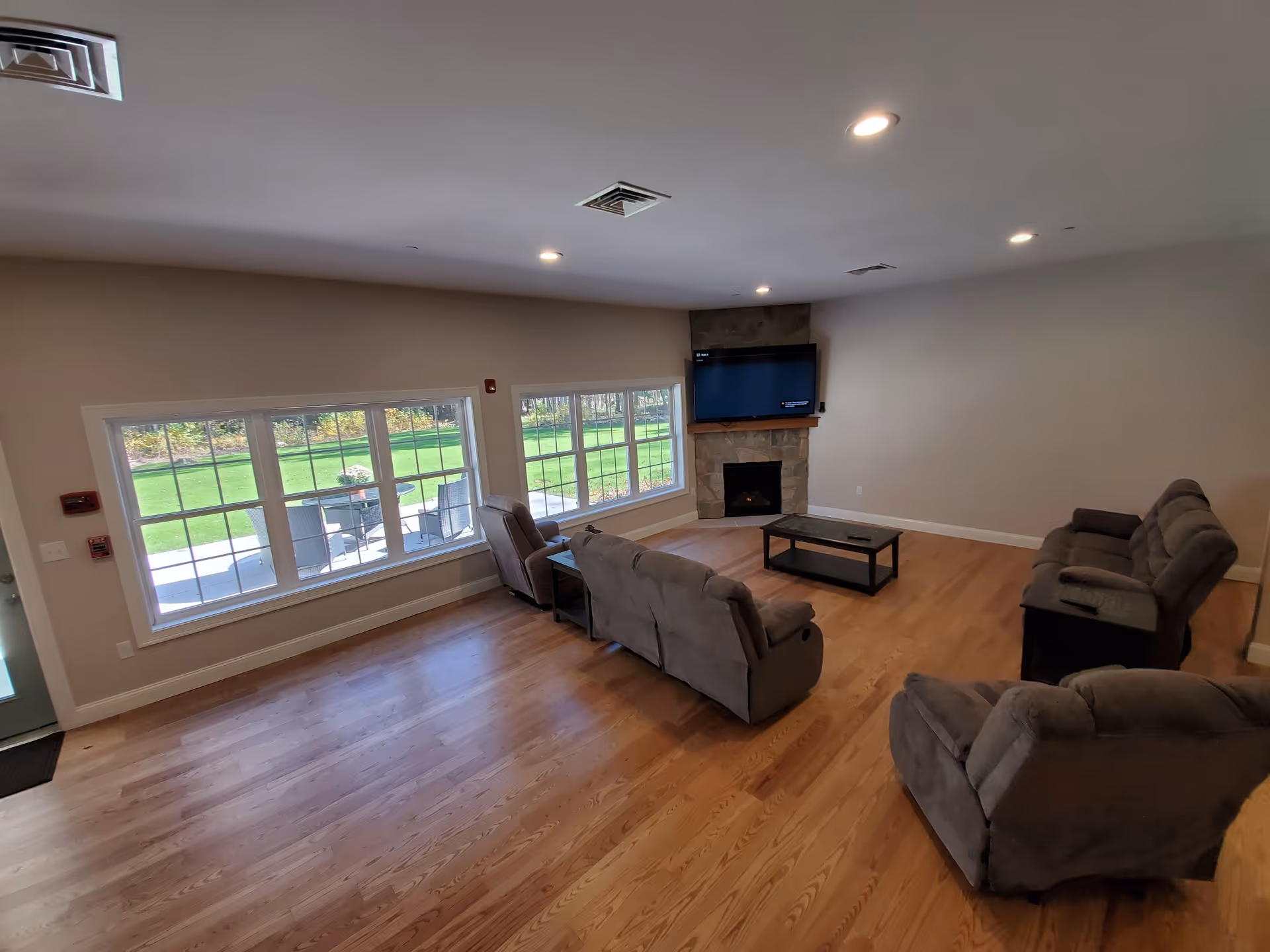 A spacious living room with wooden flooring, featuring three gray recliner sofas arranged around a black coffee table. A stone fireplace with a mounted flat-screen TV is on the far wall. Large windows on one side provide a view of a green outdoor area with patio furniture.
