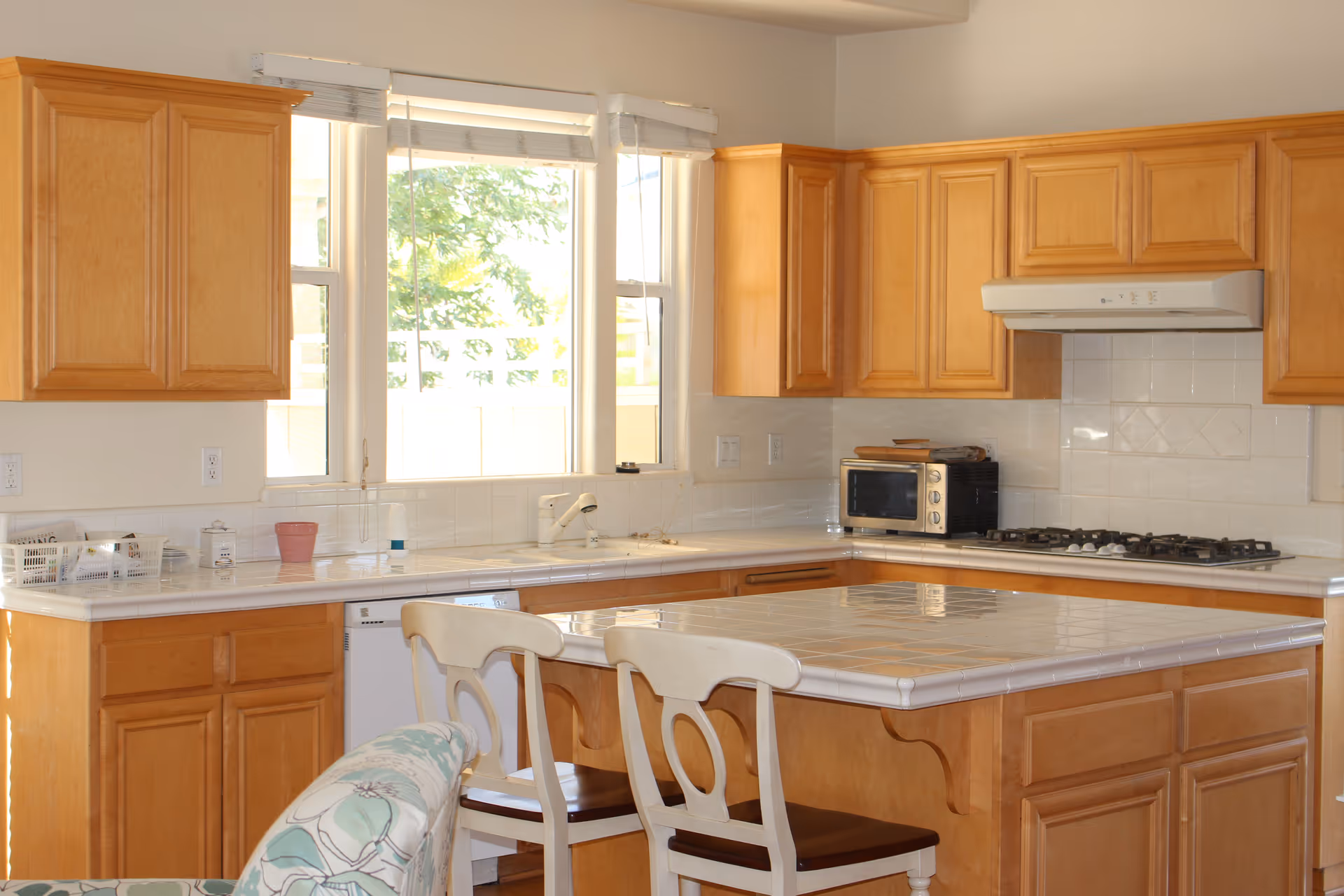Bright kitchen with wooden cabinets, a tiled island with two chairs, a gas cooktop, microwave, and a sink under a large window.