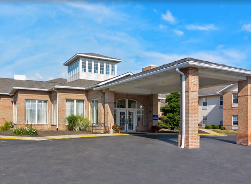 Covered portico and main entrance of a brick senior living building with large windows and a driveway under a blue sky.
