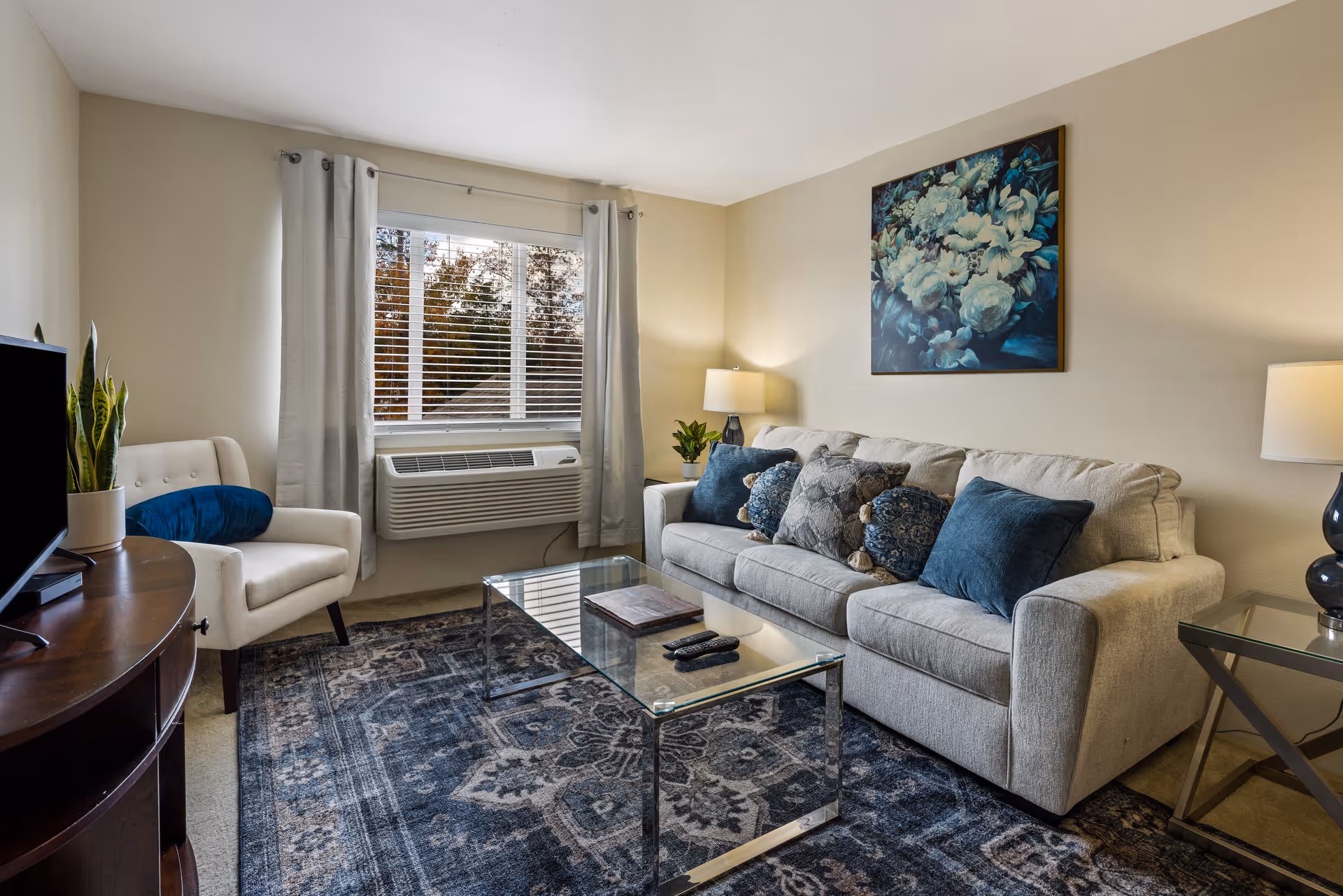A cozy living room with a beige sofa adorned with blue and patterned throw pillows, a glass coffee table, a white armchair with a blue cushion, a TV on a wooden stand, two table lamps, a large window with gray curtains, and a floral painting on the wall.