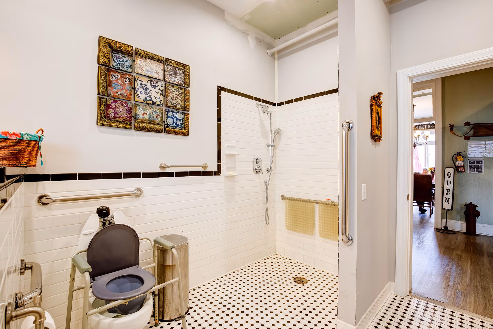 A senior-friendly bathroom with white tiled walls and black trim, featuring a walk-in shower with grab bars and a handheld showerhead. There is a raised toilet with armrests and a trash can beside it. A colorful framed artwork hangs on the wall above the toilet. The bathroom floor has a black and white hexagonal tile pattern. The door opens to a hallway with wooden flooring and a sign that reads 'OPEN'.
