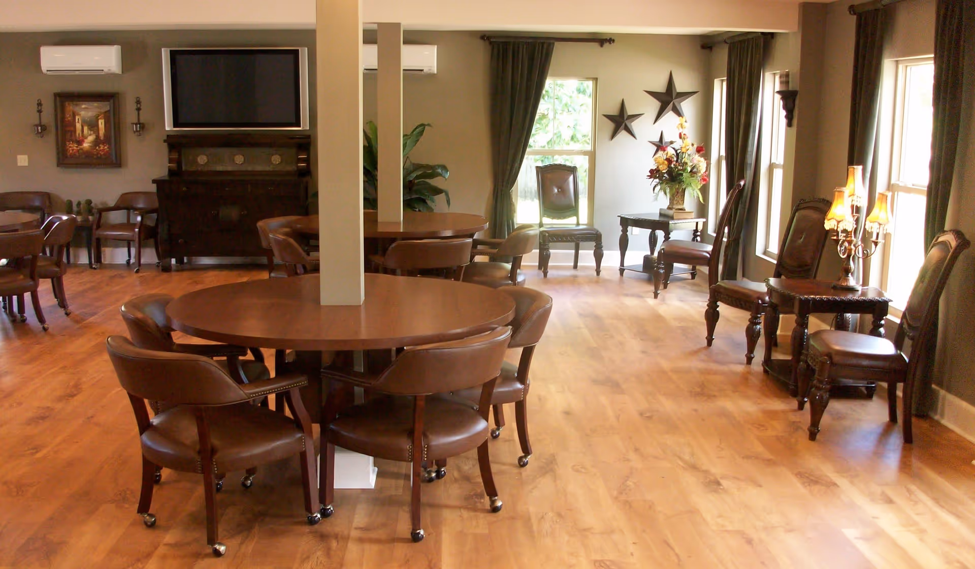 A spacious room with wooden floors and multiple round wooden tables surrounded by brown leather chairs on casters. The room has large windows with dark curtains, decorative wall stars, a floral arrangement on a small table, and a flat-screen TV mounted on the wall above a dark wooden cabinet. The lighting is warm and inviting.