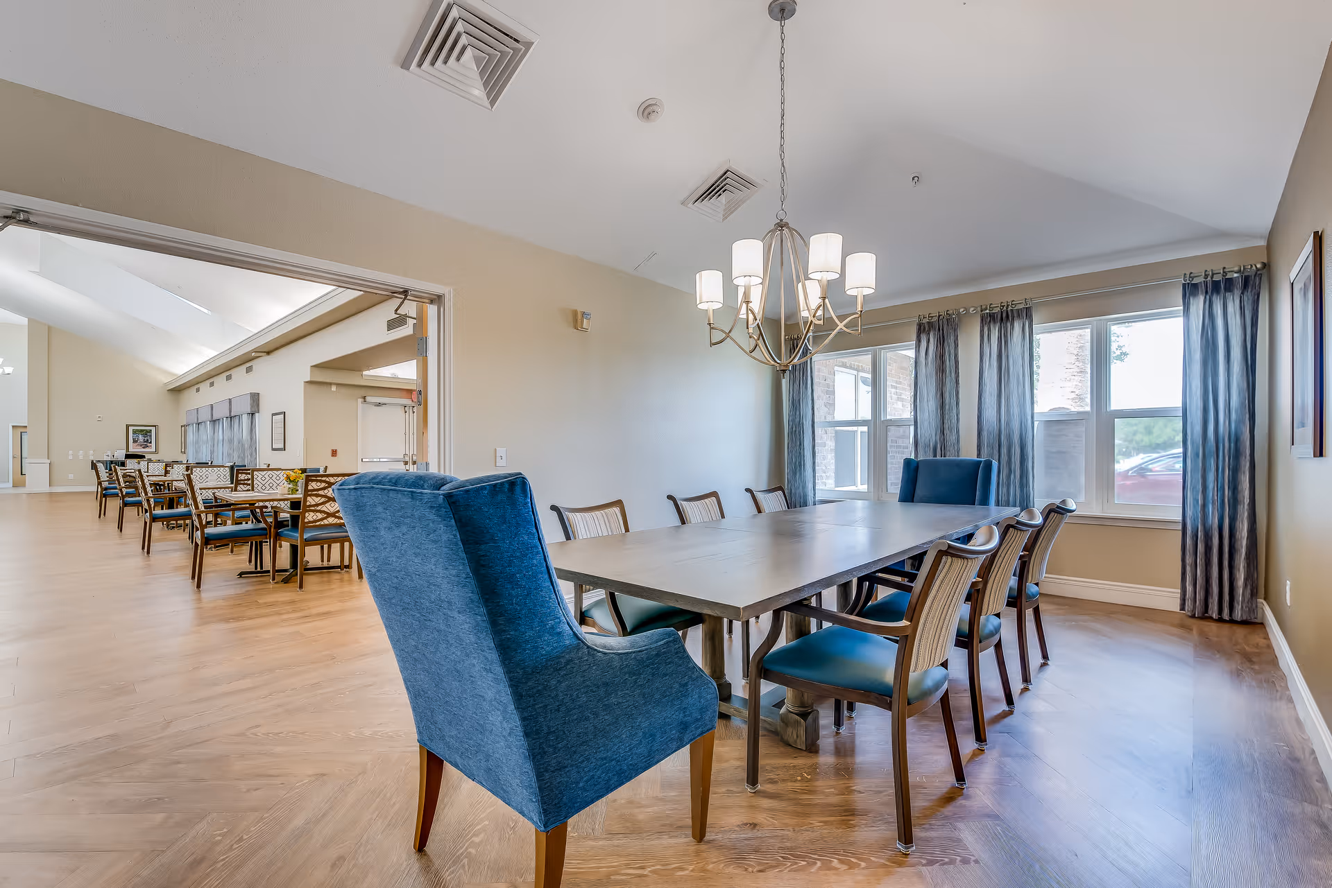 A spacious dining room with a long rectangular table surrounded by chairs, including two blue upholstered armchairs at the ends. The room has large windows with blue curtains allowing natural light to enter. A modern chandelier hangs above the table. In the background, there is an adjoining room with more tables and chairs arranged in rows.