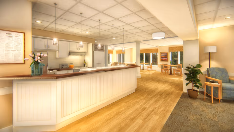 A bright and spacious interior area featuring a long kitchen counter with white cabinetry and pendant lights hanging above. The kitchen area includes appliances such as a refrigerator and stove. Beyond the counter, there is a dining area with several tables and chairs near large windows letting in natural light. To the right, there is a cozy seating area with an armchair, a small side table, a floor lamp, and a potted plant. The flooring transitions from wood in the kitchen and dining area to carpet in the seating area.