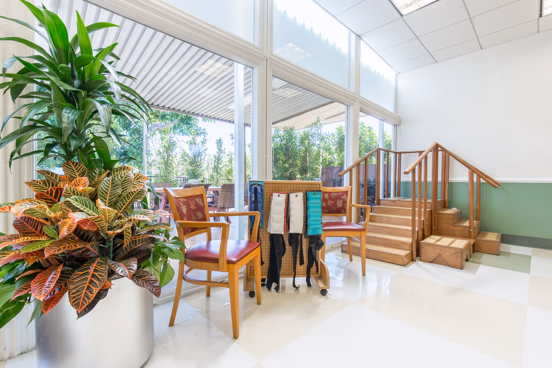 Bright indoor area with large windows showing an outdoor patio. Two wooden chairs with red cushions are placed near a pegboard holding various exercise bands. There is a large potted plant with green and orange leaves on the left side. Wooden stairs with handrails lead to a small elevated platform on the right. The floor is tiled in white and light green.