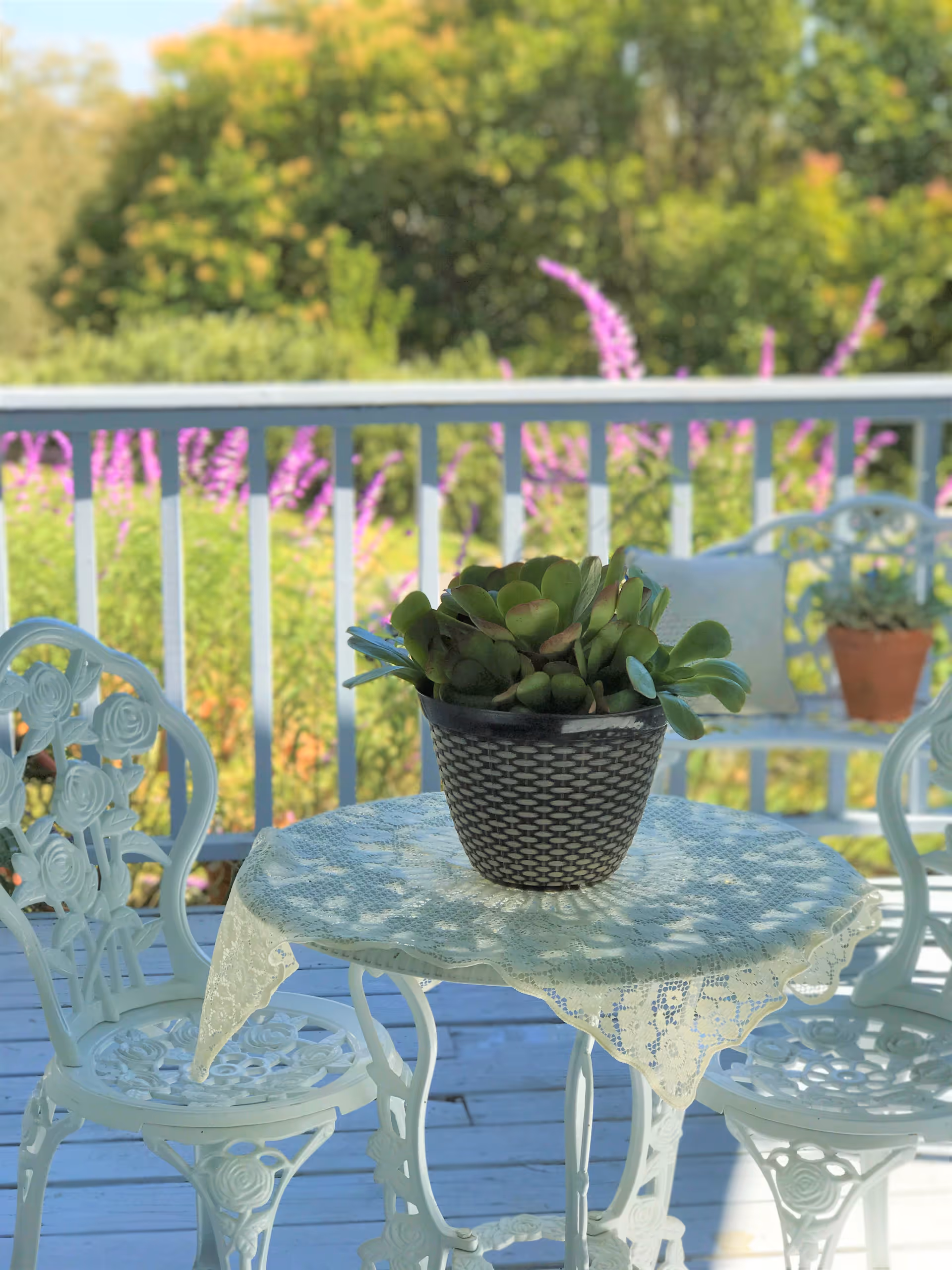 White metal patio table and chairs on a porch with a potted succulent on a lace tablecloth and flowering garden beyond the railing.