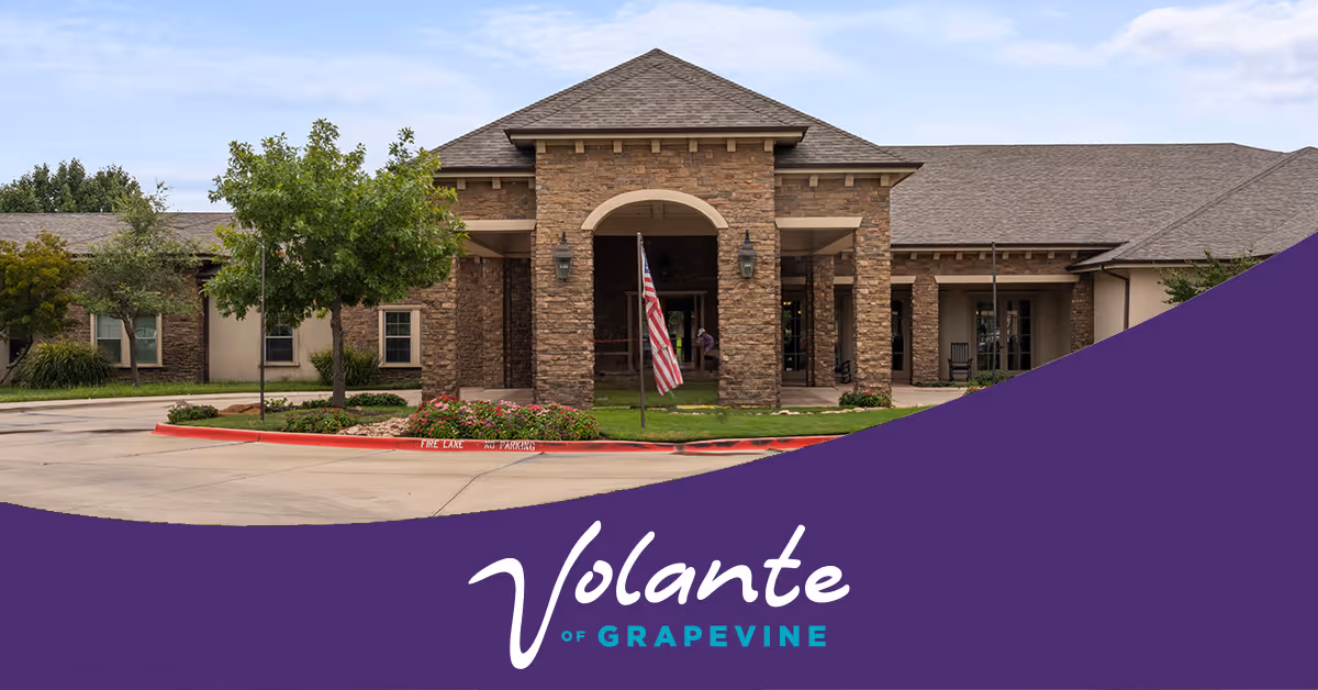 Front exterior view of Volante Senior Living of Grapevine, showing a stone and brick building entrance with an American flag in front, surrounded by trees and landscaping.