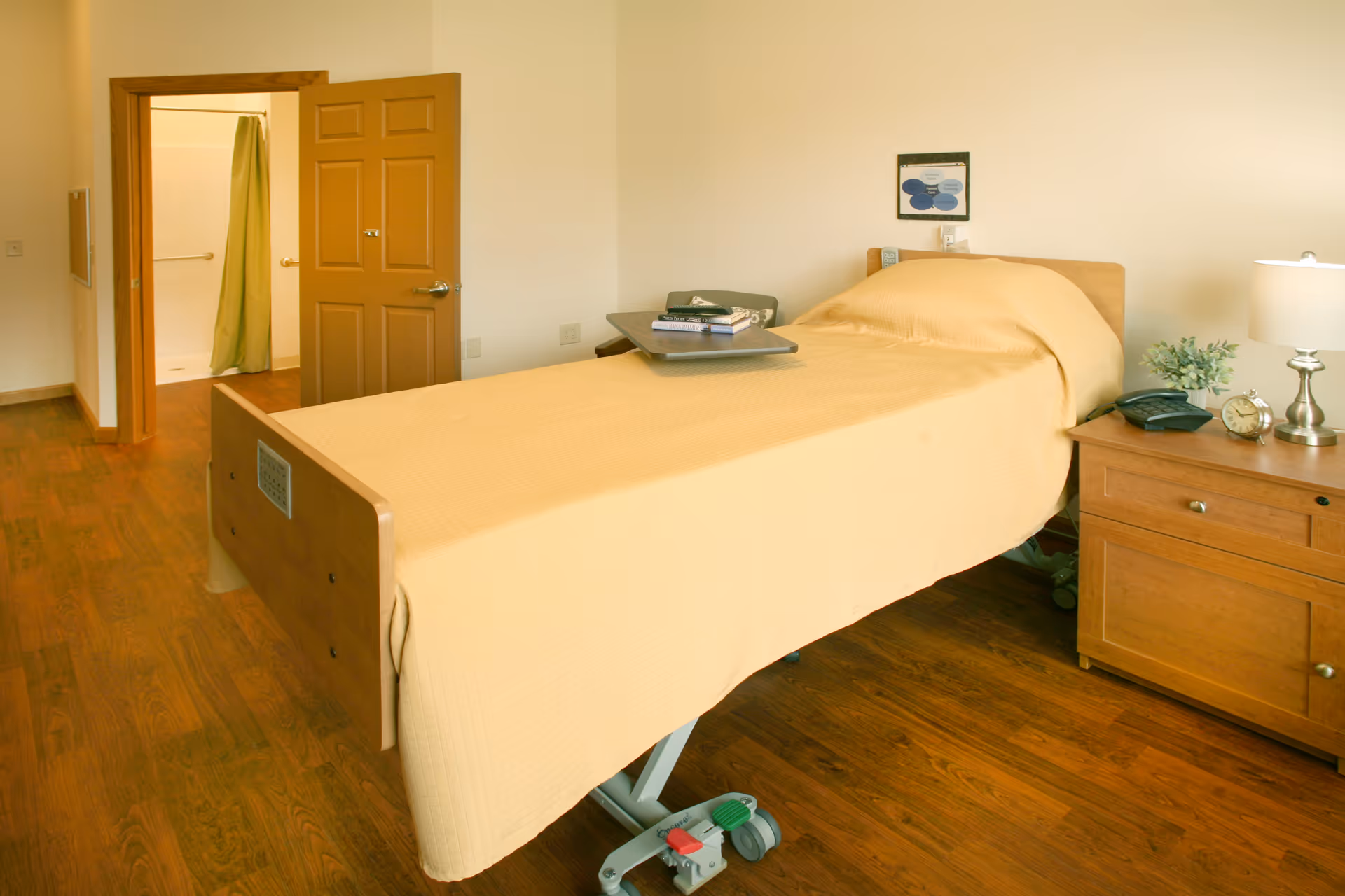 A single bed with a yellow bedspread in a room with wooden flooring. There is a wooden nightstand next to the bed with a lamp, a clock, a phone, and a small plant. An open door reveals a bathroom with a shower curtain. A tray with books and a remote control is placed on the bed.