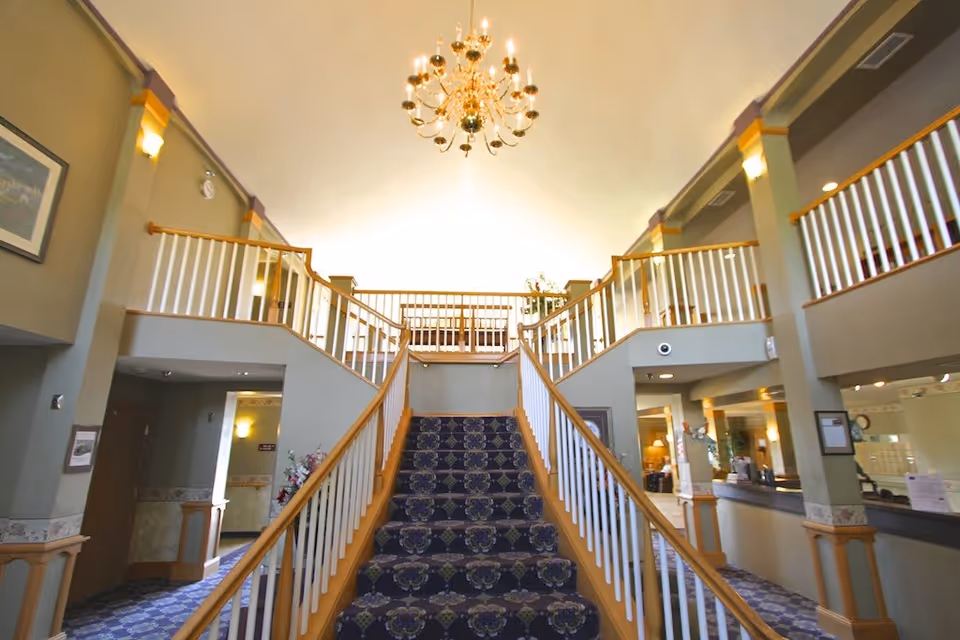 Interior view of Heathers Manor featuring a grand staircase with patterned carpet leading to an upper level with white railings. The space is well-lit with wall sconces and a central chandelier hanging from the ceiling. There are seating areas and a reception desk visible on the right side.