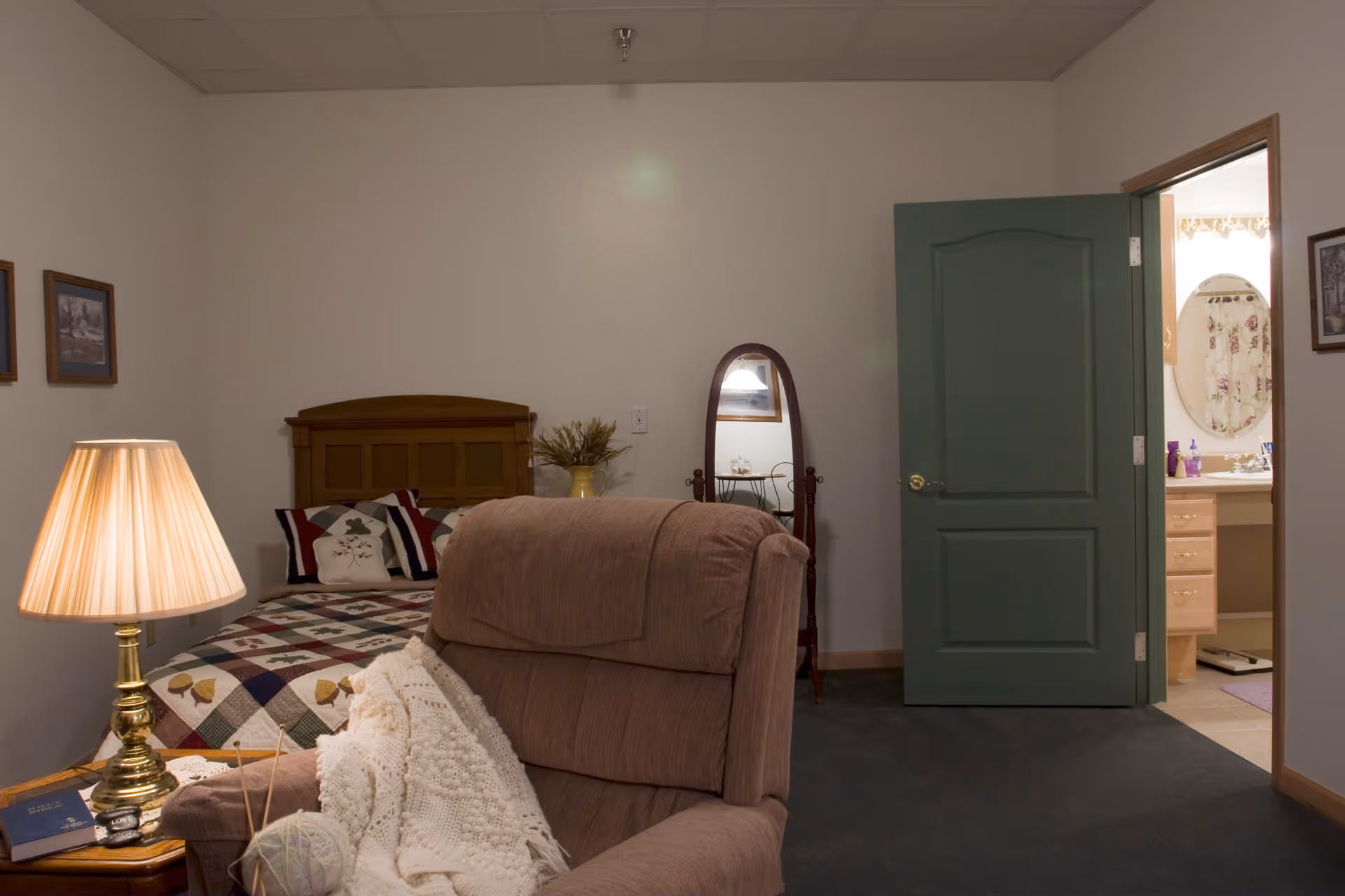 A cozy bedroom in an assisted living facility featuring a wooden bed with a patchwork quilt and decorative pillows, a beige recliner chair with a white knitted blanket and yarn, a wooden side table with a brass lamp and books, a standing oval mirror, and an open green door leading to a bathroom with a floral shower curtain and oval mirror.