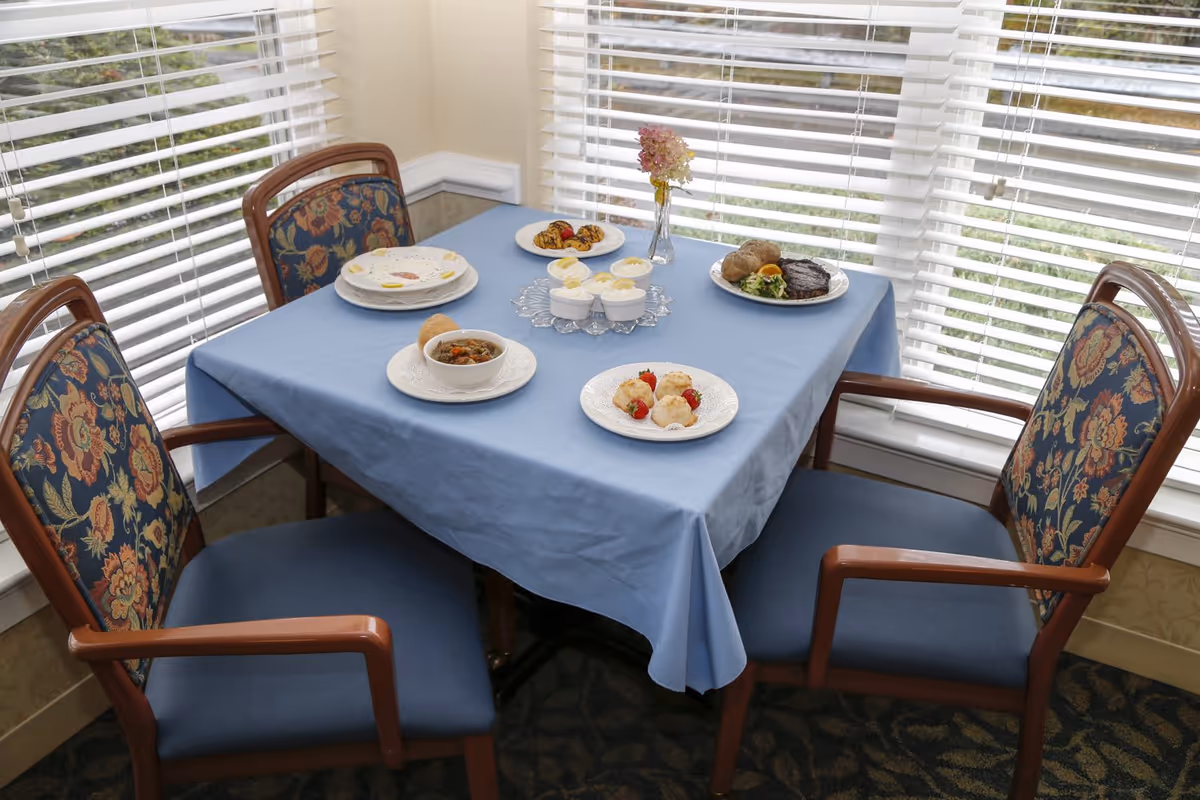 A dining table covered with a blue tablecloth set with various dishes including soup, biscuits with strawberries, cupcakes, and a plate with steak and baked potato. The table is surrounded by four wooden chairs with floral patterned cushions, and there are large windows with white blinds in the background.