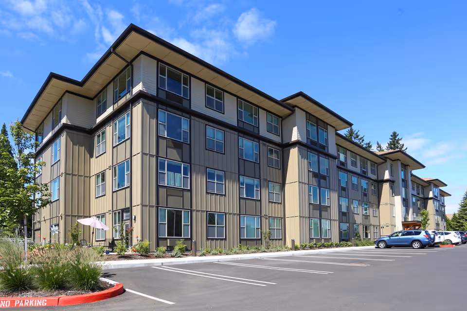 Exterior view of a modern four-story senior living facility building with multiple windows, a clear blue sky, landscaped greenery, and a parking lot with several parked cars.