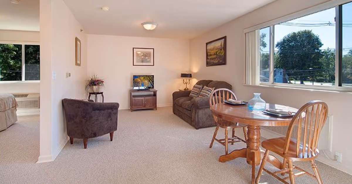 A cozy senior living room with a brown sofa, a matching armchair, a wooden round dining table with two chairs, a small TV on a wooden stand, and large windows letting in natural light. There are framed pictures on the walls and a small side table with a flower vase.
