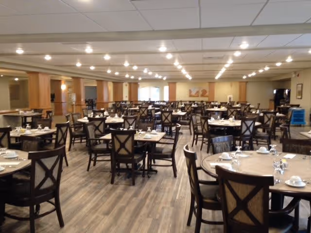 A spacious dining room in an assisted living facility with multiple round tables set with white cups, saucers, and glasses. The room has wooden flooring, beige walls, and a ceiling with recessed lighting. Dark wooden chairs with cushioned seats surround each table.