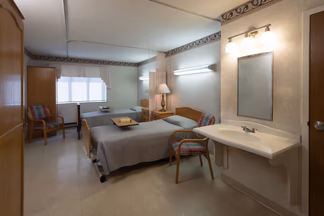 A shared senior care bedroom with two hospital-style beds, bedside tables, chairs, a wall-mounted sink and mirror, and soft overhead lighting.
