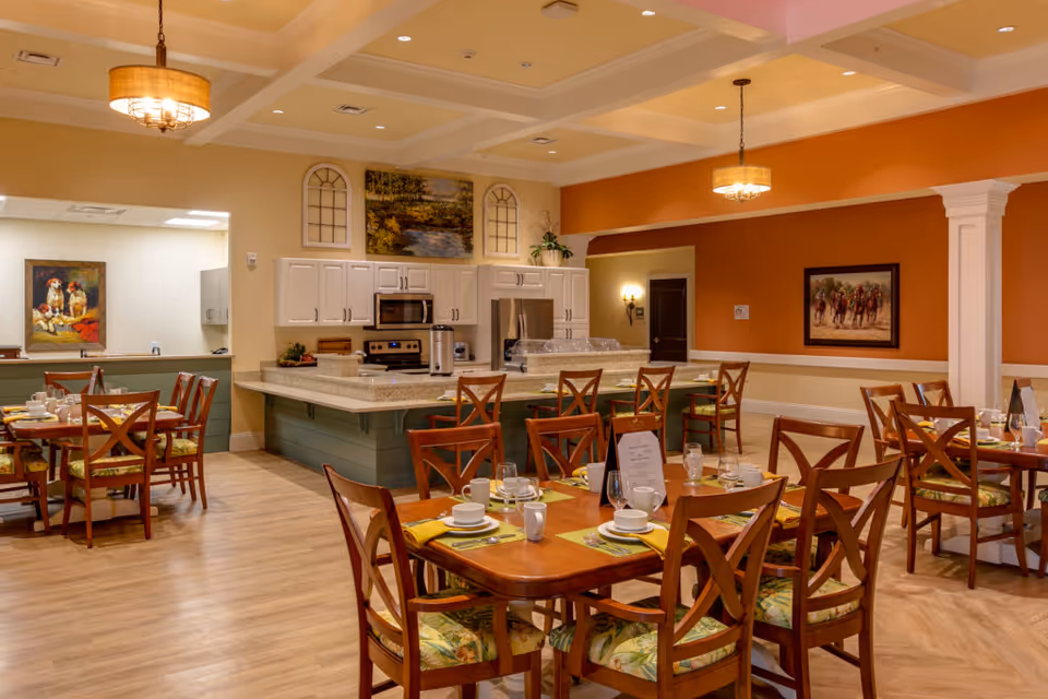 A warm and inviting dining room in a senior living facility featuring several wooden tables set with white dishes, cups, and yellow napkins. The room has wooden chairs with patterned cushions, a kitchen area with white cabinets, stainless steel appliances, and a countertop with bar stools. The walls are painted in soft beige and orange tones, decorated with framed paintings and plants. Two pendant lights hang from the ceiling, illuminating the space.