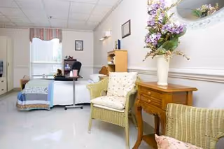 A care facility bedroom with a bed, desk, wicker chairs, and flower decor.