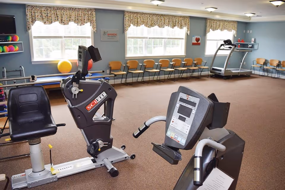 A fitness room in a senior living facility with exercise equipment including a recumbent bike and a treadmill. The room has blue walls, large windows with floral curtains, a row of wooden chairs along the walls, and colorful exercise balls on a shelf.
