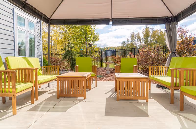 Outdoor patio area with wooden chairs and sofas featuring bright green cushions, two wooden coffee tables, a canopy overhead, and a view of trees and shrubs with a black metal fence in the background under a partly cloudy sky.