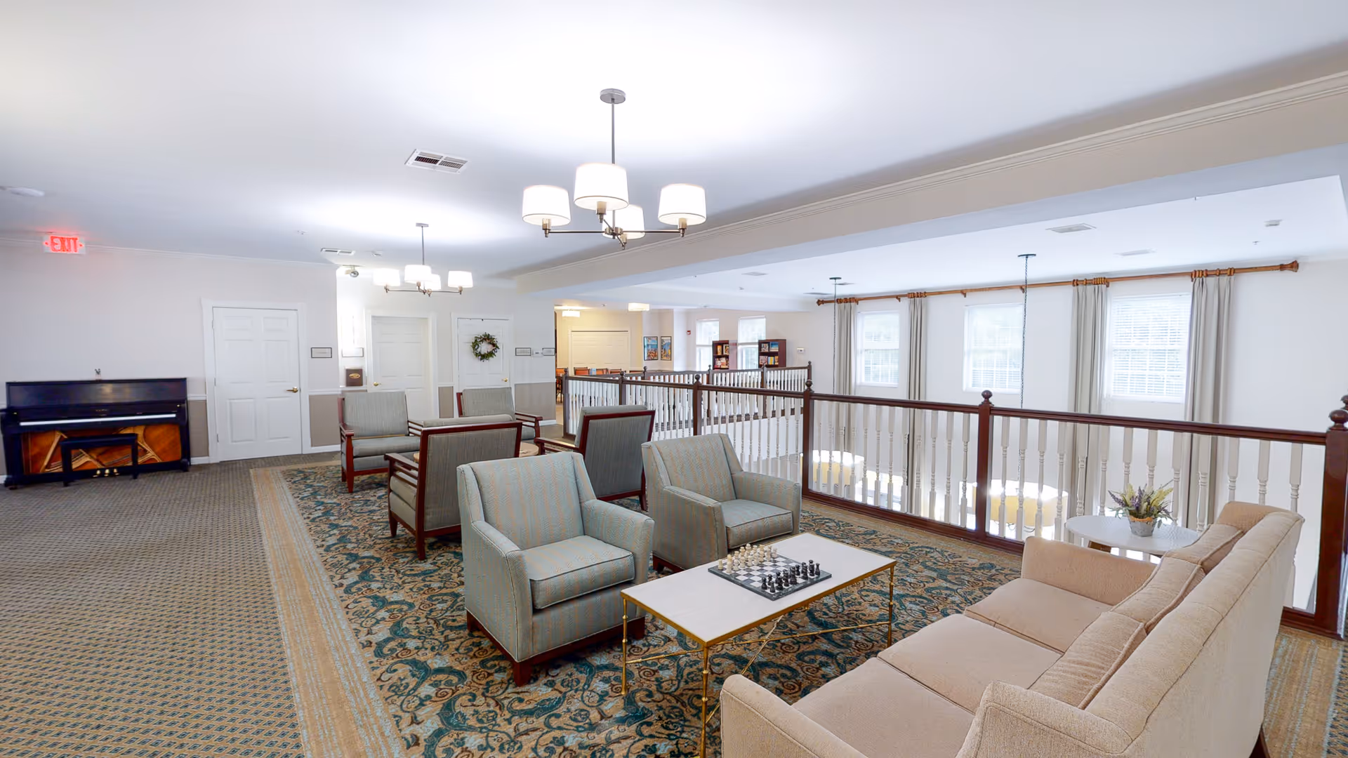 Bright communal lounge with sofas and armchairs arranged around a coffee table with a chessboard, overlooking an atrium railing.
