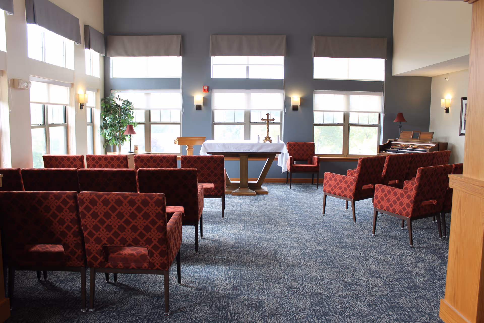 A small chapel or prayer room with rows of red patterned chairs facing a wooden altar covered with a white cloth. There is a cross on the altar, large windows with white blinds letting in natural light, a piano in the corner, and a green plant near the windows.
