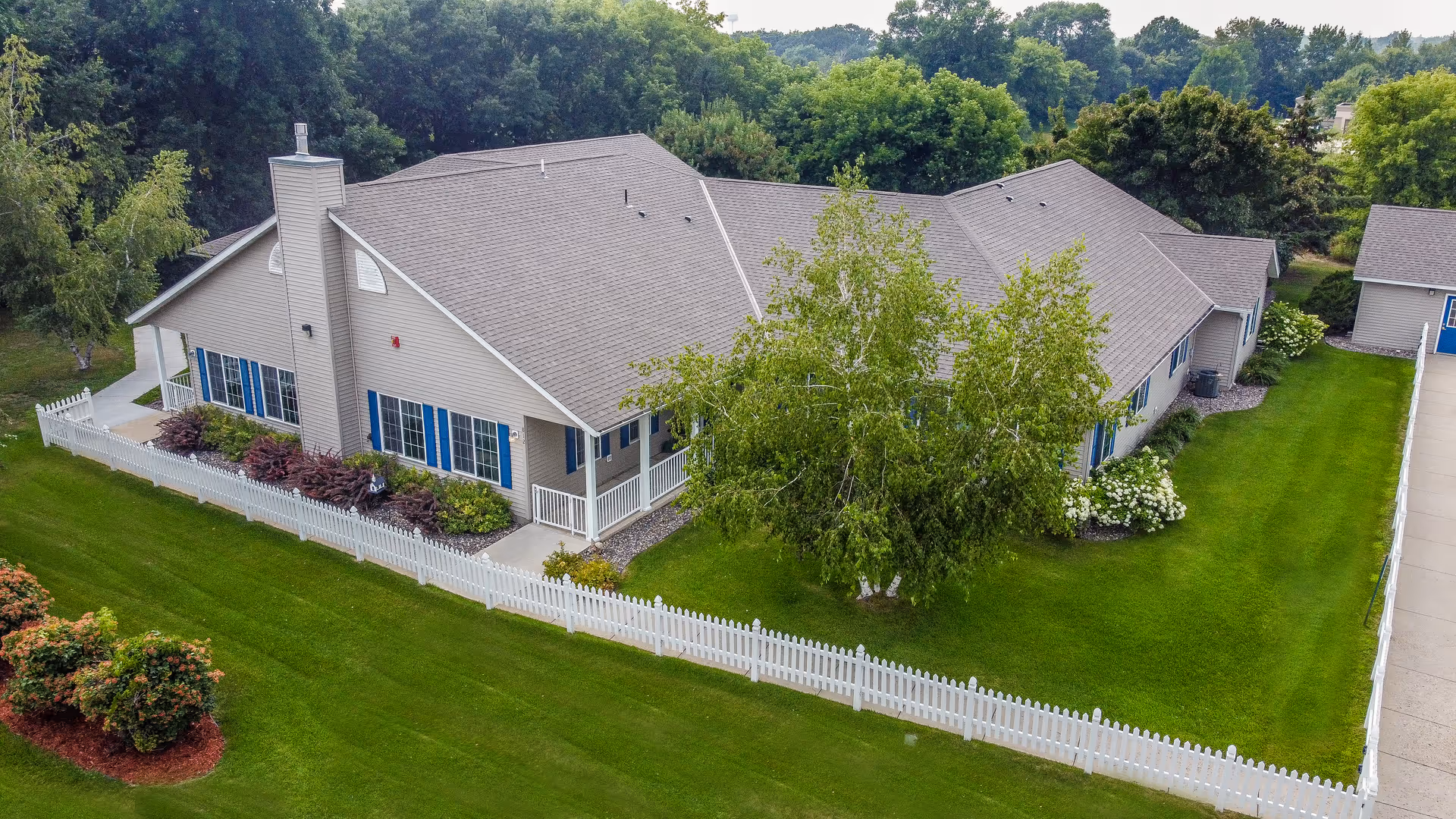 A single-story senior living building with a white picket fence, well-kept lawn, and trees surrounding it.