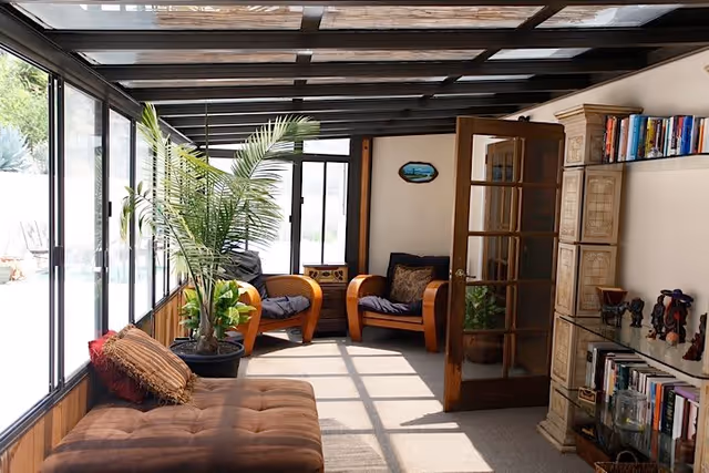 Sunlit enclosed sunroom with glass walls and ceiling beams, cushioned seating, potted plants, bookshelves and a wooden door.