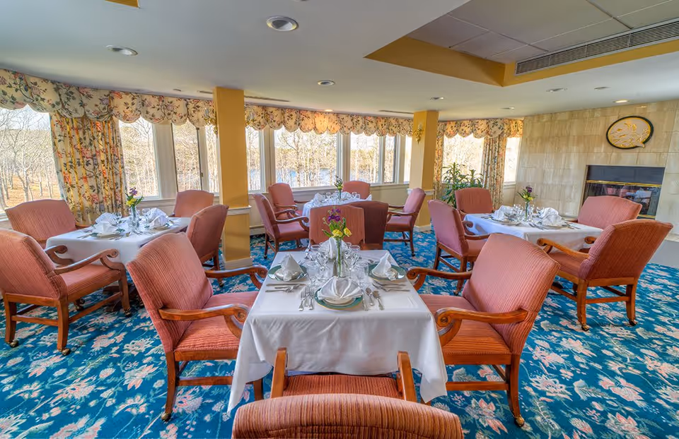 A bright dining room with multiple tables covered in white tablecloths, each set with plates, napkins, glasses, and silverware. The room features large windows with floral curtains, pink upholstered chairs, a blue floral carpet, and a fireplace on the right wall.