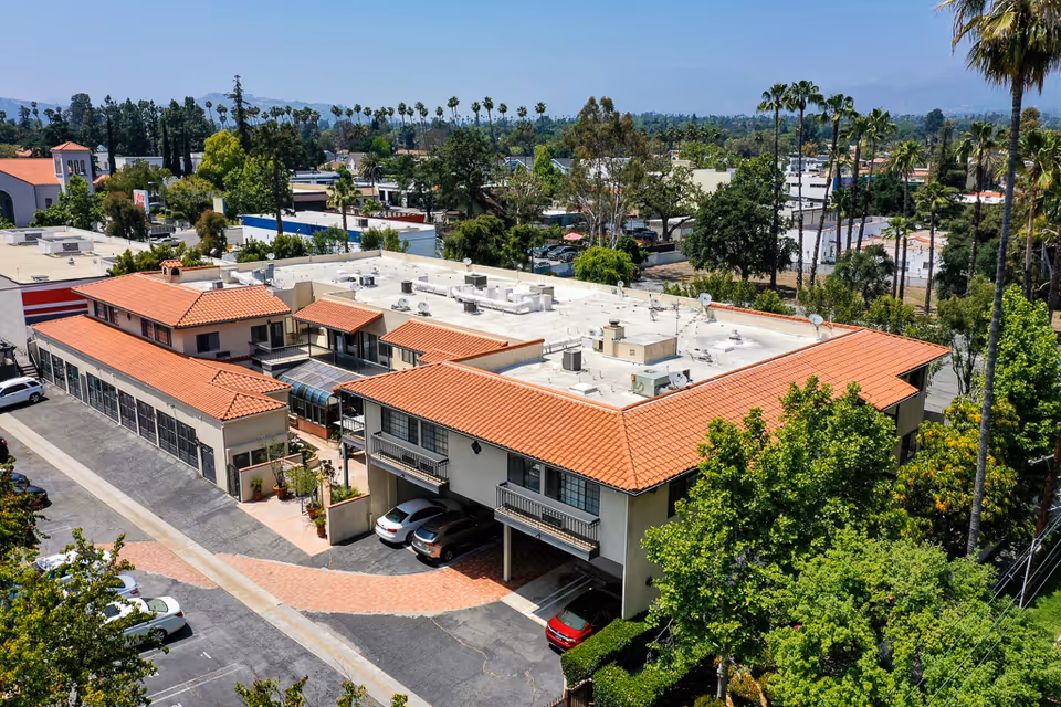 Aerial view of a senior living facility building with a red-tiled roof surrounded by trees and parking spaces, located in a suburban area with mountains in the background.