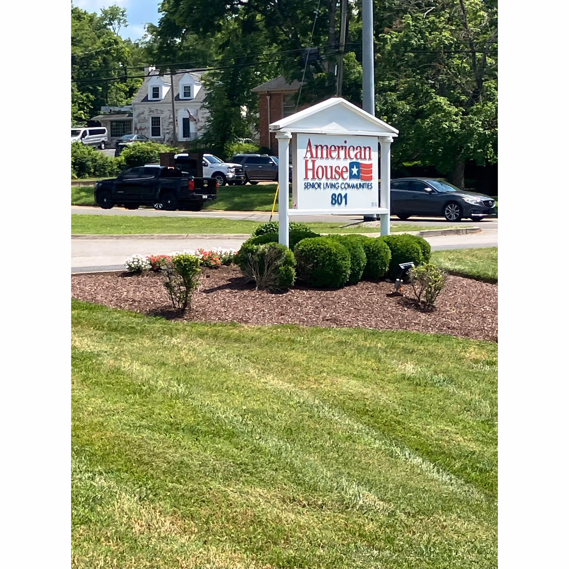 A landscaped lawn and entrance sign reading "American House Senior Living Communities 801" with parked cars and houses in the background.